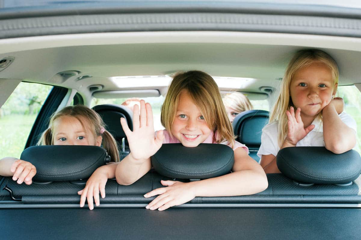 Young girls in the back of a vehicle turned around and waving out the back window.