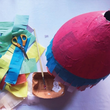 DIY paper maché piñata in progress with strips of tissue paper and a paintbrush with glue beside it.