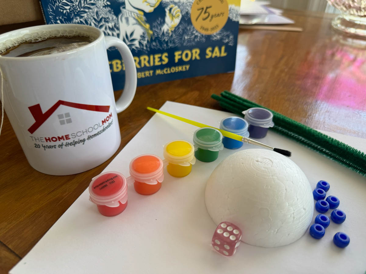 Supplies including paint, pipe cleaners, styrofoam, blue beads, and more to make a blueberry bush craft with the book Blueberries for Sal in the background.