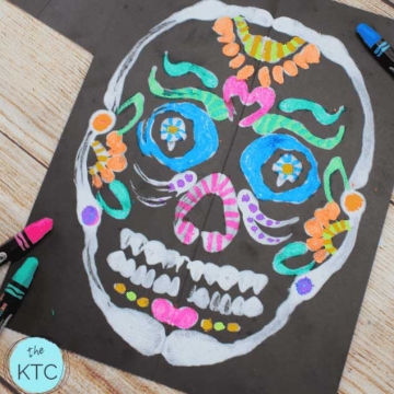 A painting of a sugar skull on black paper with white and bright color paint.