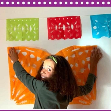 Child holding papel picado banner up to a wall beneath a streamer with multiple papel picado cut-outs in bright colors.