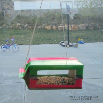 Bird feeder made from a half gallon cardboard milk container hanging at a window.